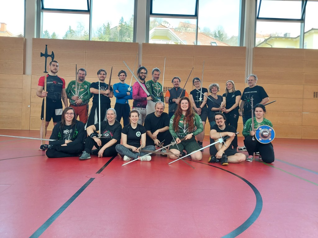 Eine Gruppe Menschen mit Trainingsschwertern, Helebarde, Schwert und Buckler in einer Sporthalle.